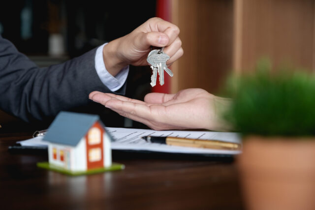 Man handing over a set of keys to former spouse across a table with financial documents on it and a small house shaped bust; Broward County Marital Property Lawyer concept image